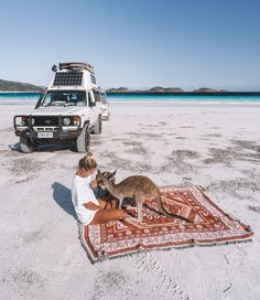 a woman sitting on top of a rug next to a kangaroo in front of an suv