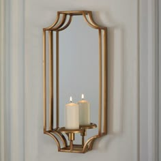 two white candles are lit in front of a mirror on the wall with gold trim