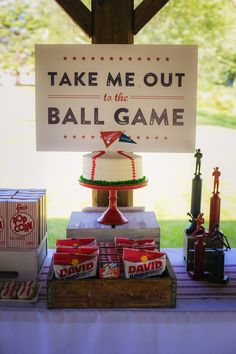 a table topped with a cake covered in red, white and blue frosting next to a sign that says take me out to the ball game