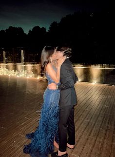 a man and woman kissing on a wooden deck at night with lights in the background