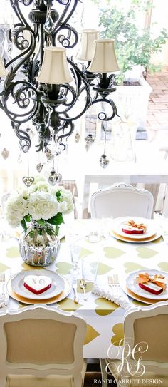 a chandelier hanging over a dining room table with plates and flowers on it