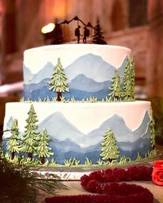 a three tiered cake with trees and mountains painted on the side, surrounded by greenery