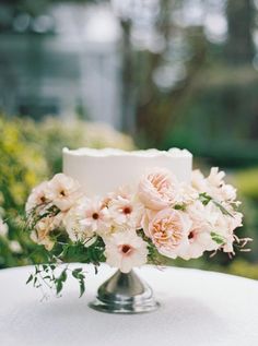 a white wedding cake with pink flowers on the top is sitting on a table outside