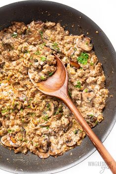 a skillet filled with mushroom and meat mixture next to a wooden spoon on a white surface