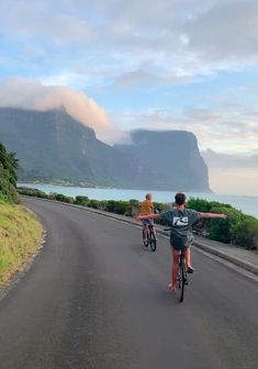 two people riding bikes down the road with mountains in the background