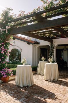 two tables covered with white tablecloths under an outdoor pergolated area in front of a house