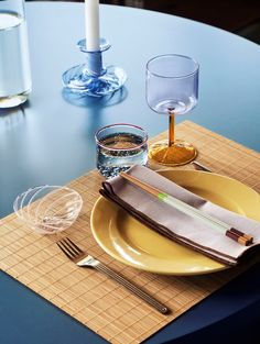 a place setting with yellow plates and silverware on a blue table cloth, next to a lit candle