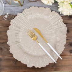 an empty plate with two gold forks on it next to some wine glasses and flowers