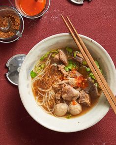 a white bowl filled with meat and noodles next to chopsticks on a red table