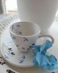 a cup and saucer on a plate with a blue flower next to it in front of a white vase