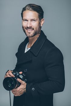 a man holding a camera and smiling at the camera while wearing a black coat with a white t - shirt