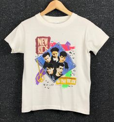 "Tag Size: Children's 10-12 - Fit: See measurements below - Front: Band image - multi-color on white - Reverse: none - 100% cotton - Short sleeves - Crew neckline - Single-stitching Condition: Good / vintage - Copyright 1990 Step Productions (pictured) APPROXIMATE MEASUREMENTS - Pit-Pit: 16 1/2\" - Shoulder: 14 1/2\" - Length: 20 1/2" 90s Style White Shirt With Screen Print, Retro White Tops With Band Logo, White 90s Style Top For Concerts, Multicolor Cotton Top For Concerts, White Unisex Tops With Band Logo, Pop Culture Cotton Top With Band Logo, White Unisex Top With Band Logo, Retro White Top With Band Logo, White 90s T-shirt For Concert