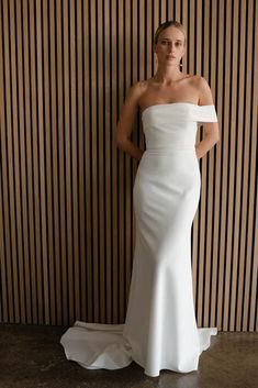 a woman standing in front of a striped wall wearing a white wedding dress with an off the shoulder neckline