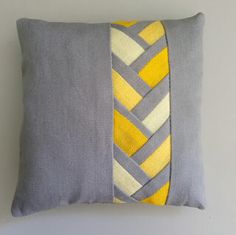 a gray and yellow pillow with braiding on the front, sitting against a white wall