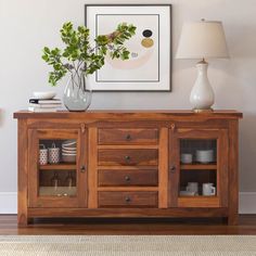 a wooden sideboard with glass doors and two vases on top, next to a lamp