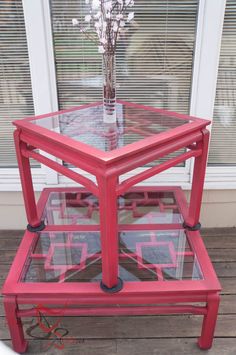 a red table with glass top and flowers in a vase on the bottom, next to a window