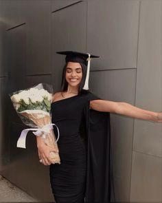 a woman in a graduation cap and gown holding a bouquet of flowers while standing next to a wall