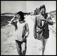 an old black and white photo of two people walking on the beach, one holding a cell phone to his ear