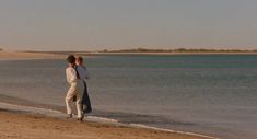 two people walking on the beach next to the water