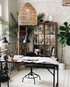 an office with plants and books on the desk, in front of a window that has a wicker hanging over it