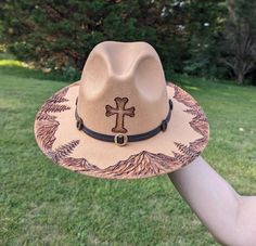 This unique fedora makes a great addition to any wardrobe. The warm tan material burns a rich deep brown allowing the design to stand out. A cross is intricately burned and shaded on the crown by hand using a pyrography machine. A detailed landscape of a mountain range and pine trees is sculpted and shaded along the entire circumference of the brim. All my hat designs are completely original and freehanded. For a beautiful finishing touch the hat is embellished with a western faux leather hat band.  Hat Materials: Cotton and polyester Hat fits most women with an adjustable tie band inside *Disclaimer* Colors may vary slightly screen to screen Rustic Brown Brimmed Panama Hat, Rustic Brown Fedora With Curved Brim, Rustic Brown Panama Hat With Curved Brim, Rustic Brown Flat Brim Fedora, Brown Curved Brim Panama Hat For Western-themed Events, Brown Fedora With Short Brim For Country Events, Rustic Brown Brimmed Fedora, Brown Sun Hat For Western-themed Events With Short Brim, Custom Brown Fedora With Curved Brim