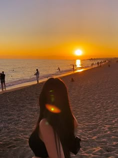 a woman sitting on top of a sandy beach next to the ocean at sun set