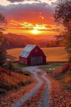 a red barn sits on the side of a dirt road as the sun sets behind it