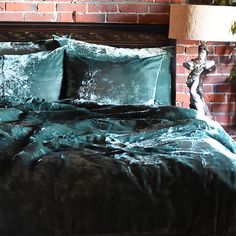 a bed with green sheets and pillows in front of a brick wall, next to a lamp