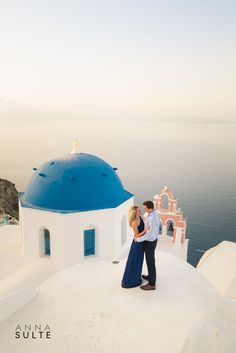 a man and woman standing on top of a white building next to the ocean with blue domes