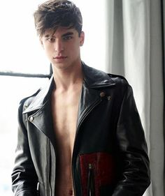 a shirtless man in a leather jacket standing by a window with his hands on his hips