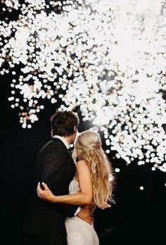 a bride and groom standing in front of a fireworks display with their arms around each other