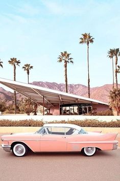 an old pink car parked in front of a building with palm trees and mountains in the background