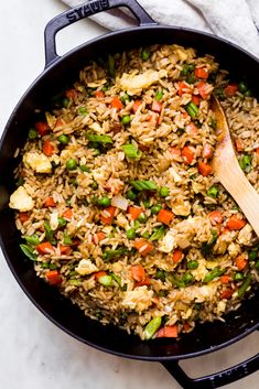 a skillet filled with rice and vegetables on top of a white countertop next to a wooden spoon