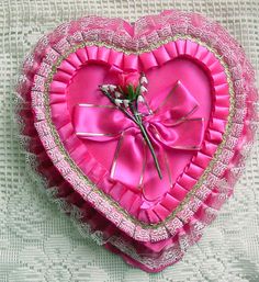 a pink heart shaped box sitting on top of a white cloth covered tablecloth with a flower in the center