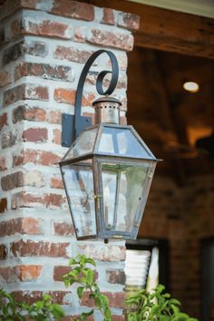 an old fashioned light hanging from the side of a brick building with green plants in front of it