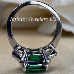 "-Approximate total carat weight: approx. 6.00ctw diamond equivalent #7080 -Center Stone Size: 11x9mm - approx. 4.90ct diamond equivalent -Center Stone Shape: emerald cut -Gem Type: lab created Green Emerald -Stone Clarity: VS2 -Stone Color: F -Moh's Scale: 9 hardness -Side Stones Size: 6x4mm trapezoid approx. 0.55ct. each diamond equivalent -Gem Type: Zonar moissanite -Metal Type and Purity: 14k white gold -Setting: 4 prong tulip setting -Stock Ring Size: 5.75 -Country of Manufacturing: USA (Mi White Gold Tsavorite Anniversary Jewelry, White Gold Tsavorite Emerald Ring For Anniversary, Platinum Emerald Three Stone Ring For Anniversary, White Gold Tsavorite Jewelry For Anniversary, Platinum Three Stone Emerald Anniversary Ring, Anniversary Fine Jewelry Tsavorite Rings, Gia Certified Green Princess Cut Rings, Platinum Green Ring For Anniversary, Green Platinum Anniversary Rings