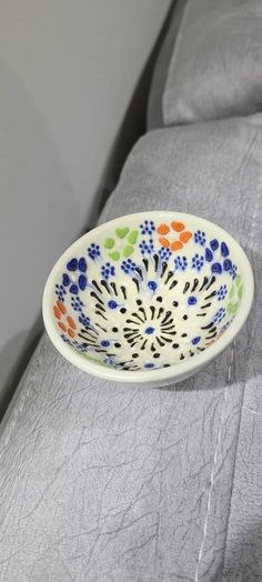 a white bowl with blue, orange and green designs on it sitting on a gray couch