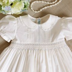 This Ivory Smocked Heirloom Dress By Feltman Brothers Is Darling! The Traditional Style Is Timeless And Super Classy. The Ivory Color Is Very Classy And A Lovely Change From Basic White. The Cotton Blend Dress Has A Peter Pan Collar Trimmed In A Delicate French Lace. The Bodice Has Vertical Pin-Tucks With Light Blue Feather Stitched Embroidery In The Center. Just Below The Pin Tuck Detail Is A Matching Piping Bordering A Smocked Was It. The Smocking Has A White And Blue Embroidered Lattice Design. The White Is A Lovely Accent That Ties In With The Lace On The Collar. The Sleeves Are Short And Puffed With A Matching Half Inch Band. Upper Button Back. A Tie-Back Sash For The Perfect Fit O Heirloom Baby Clothes, Feltman Brothers, Baby Heirloom, Floral Ruffle Dress, Couture Sewing Techniques, Cotton Blends Dress