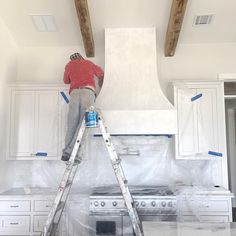 a man standing on a ladder painting the ceiling in a kitchen with white cabinets and wood beams