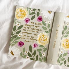 an open book with flowers on it sitting on top of a white bed sheet next to another book