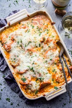 a casserole dish with meat, cheese and parsley in it next to two wine glasses