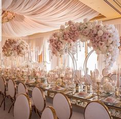 a long table with white and pink flowers on it, surrounded by gold chairs in front of a window