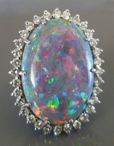 This stunning marked natural Queensland boulder opal is set in platinum and surrounded by diamonds. The oval cabochon-cut opal measures 28.00 mm x 18.75 mm (5.84 mm depth) and weighs approximately 16.34 carats. Surrounding the opal are thirty-two round full-cut diamonds, each weighing approximately 0.04 carats for an approximate total diamond weight of 1.36 carats. These high quality diamonds have a clarity grade of VS1 to VS2 and a color grade of G. What makes this large opal particularly strik Opal Ring Vintage, Opal And Diamond Ring, Boulder Opal Ring, Opal Diamond Ring, Blue Opal Ring, Ruby Diamond Rings, Garnet Earrings, Opal Ring, Gems Jewelry