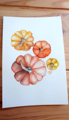 a watercolor painting of three pumpkins on a white paper with a wooden table in the background