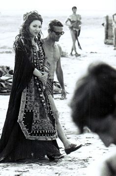 a man and woman are walking on the beach with other people in the water behind them