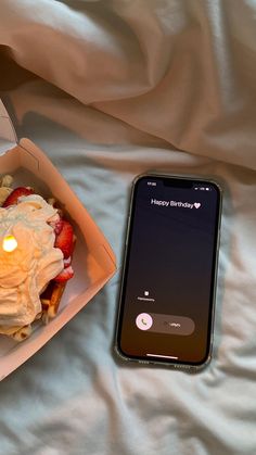 a cell phone sitting next to an open box of food on a bed with white sheets