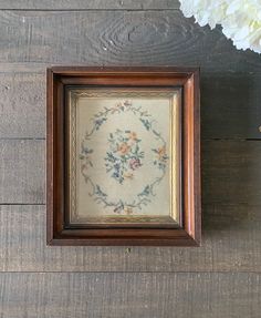 a cross stitch pattern in a wooden frame with flowers on the table next to it