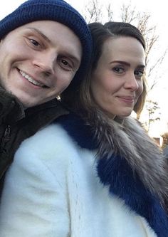 a man and woman standing next to each other in front of a building with snow on the ground