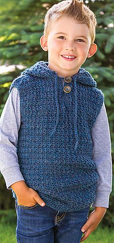 a young boy wearing a blue knitted vest and smiling at the camera with his hands in his pockets