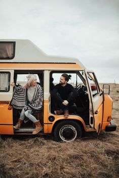 two people sitting in the open door of an orange and white van on dry grass